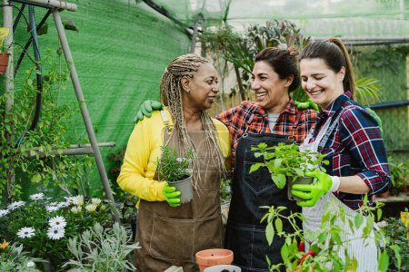 Téléchargez les photos : Heureuses femmes multiraciales travaillant ensemble dans les plantes et les fleurs magasin de jardin - en image libre de droit