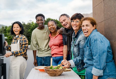 Happy multigenerational friends having fun preparing food at house rooftop - Diversity people lifestyle