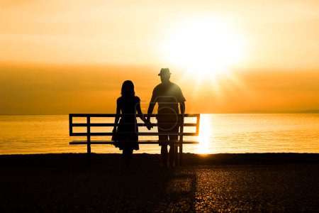 Téléchargez les photos : Couple heureux sur un banc au bord de la mer sur la nature en silhouette de voyage - en image libre de droit