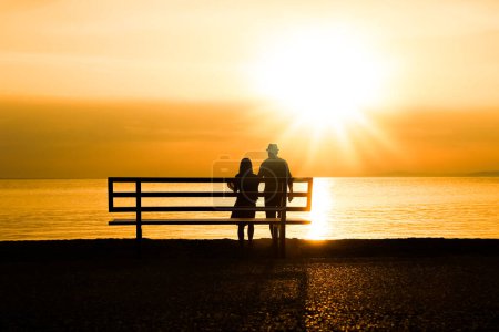 Téléchargez les photos : Couple heureux sur un banc au bord de la mer sur la nature en silhouette de voyage - en image libre de droit