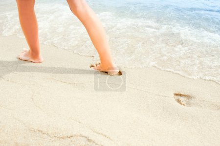 Foto de Viaje a la playa - mujer relajada caminando en una playa de arena dejando huellas en la arena. El detalle cercano de los pies femeninos sobre la arena dorada en la playa en Grecia. Contexto. - Imagen libre de derechos