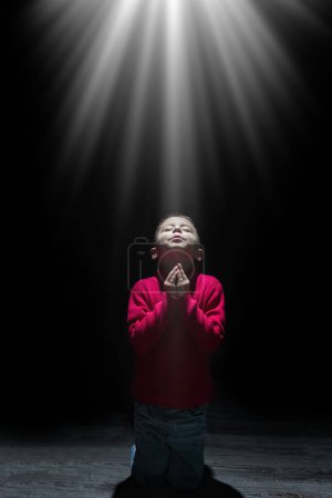 Téléchargez les photos : Enfant priant spirituellement Dieu sur un fond noir - en image libre de droit