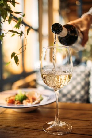 Photo for White wine poured into a glass with a served dish on the background at the restaurant - Royalty Free Image
