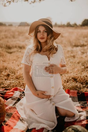 Photo for Happy European caucasian pregnant woman relaxing in nature picnic looking at camera. expectant mother in hat and dress hold hand on belly. face with moles - Royalty Free Image