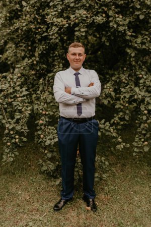 Photo for Young blond man European curly-haired successful businessman groom in classic clothes white shirt and tie stands on street with his hands clasped together in nature on background of a green leafy wall - Royalty Free Image