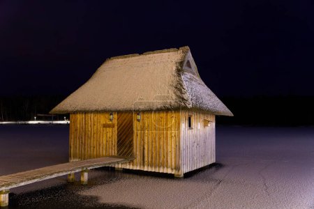 snow-covered reed roof wooden hut on a lake in the evening
