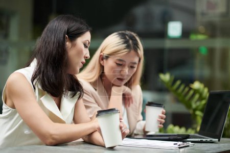 Téléchargez les photos : Pensive female entrepreneurs drinking coffee when checking marketing report at meeting - en image libre de droit