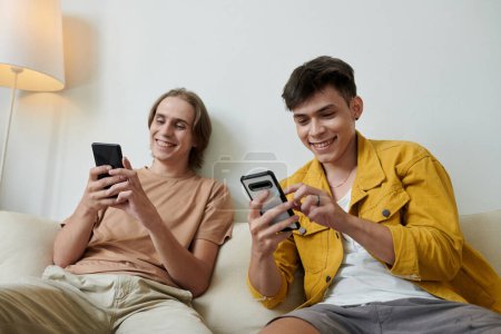 Foto de Joyful young men texting friends when sitting on couch at home - Imagen libre de derechos