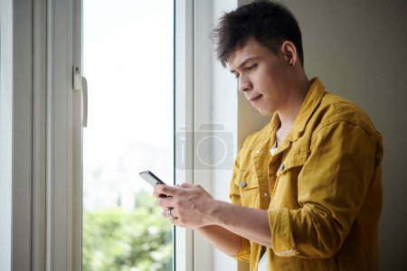 Téléchargez les photos : Young man standing at dormitory window and texting girlfriend - en image libre de droit