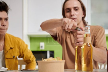 Photo for Young man opening beer bottle for himself and his friend when they are sitting at kitchen table - Royalty Free Image