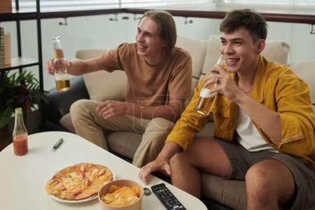 Téléchargez les photos : Happy young men drinking beer and eating pizza when watching game on tv at home - en image libre de droit