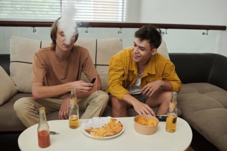 Téléchargez les photos : Young men smoking electronic cigarettes at home when having pizza with beer - en image libre de droit