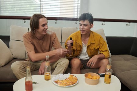 Foto de Smiling young man sharing electronic cigarette with friend when they are hanging out at home - Imagen libre de derechos