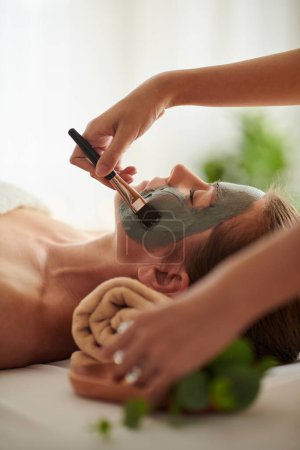 Photo for Beautician applying black clay mask on face of client in beauty salon - Royalty Free Image