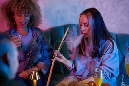 Foto de Young woman smoking hookah when spending time with friends in bar - Imagen libre de derechos