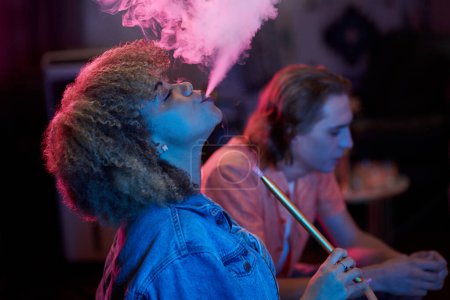 Téléchargez les photos : Side view of young Black woman exhaling hookah smoke - en image libre de droit