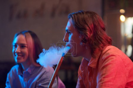 Photo for Side view of young man smoking hookah when gathering with friends in lounge bar - Royalty Free Image