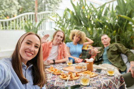 Photo for Cheerful young woman taking selfie with group of her friends when they are enjoying dinner in outdoor cafe - Royalty Free Image