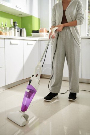 Téléchargez les photos : Housewife vacuum cleaning tile floor in kitchen - en image libre de droit