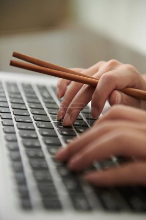 Téléchargez les photos : Hands holing chopsticks when working on laptop during lunch break - en image libre de droit