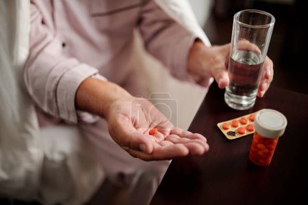 Photo for Sick elderly man sitting in bed wrapped up in comforter and taking pills - Royalty Free Image