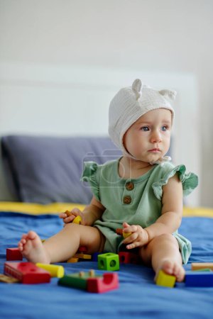 Photo for Pensive little girl sitting on bed with colorful wooden toys and looking away - Royalty Free Image