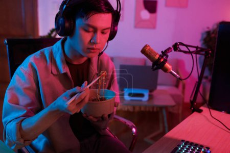 Photo for Teenage boy in headphones eating ramen and watching video of blogger on computer - Royalty Free Image