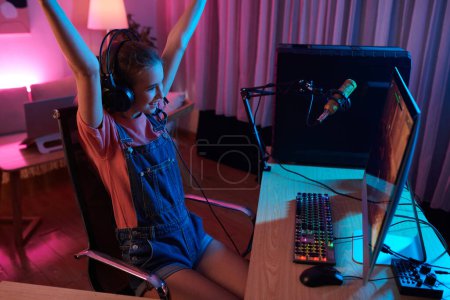 Photo for Excited teenage girl celebrating winning videogame when sitting at desk in neon bedroom - Royalty Free Image