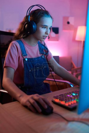 Téléchargez les photos : Teenager wearing headphones when playing videogame on computer at her desk - en image libre de droit