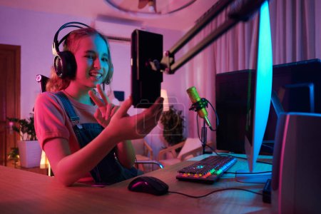 Photo for Smiling teenage girl streaming herself playing videogame and answering questions of subscribers - Royalty Free Image