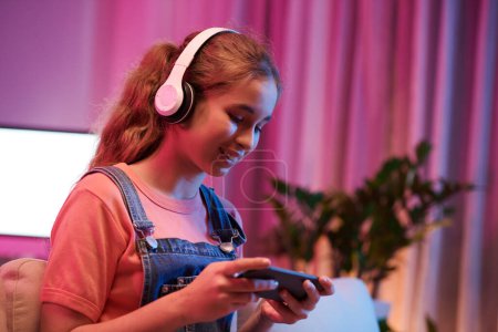 Foto de Happy teenage girl in headphones watching entertaining videos on smartphone - Imagen libre de derechos