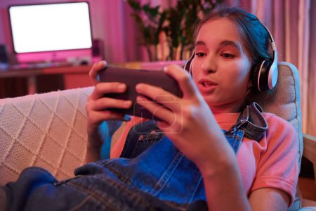 Foto de Excited teenage girl relaxing on sofa and watching videos of her favorite blooger on smartphone - Imagen libre de derechos