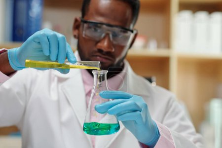 Foto de Laboratory worker in protective glasses mixing green and yellow liquids - Imagen libre de derechos