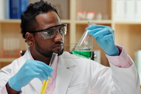 Foto de Serious researcher looking at flask and test-tube with green and yellow liquids - Imagen libre de derechos