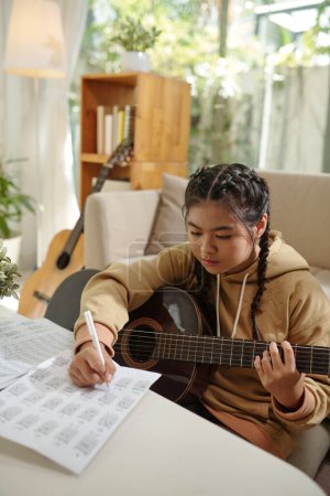 Photo for Concentrated teenage girl writing down music notes when playing guitar at home - Royalty Free Image