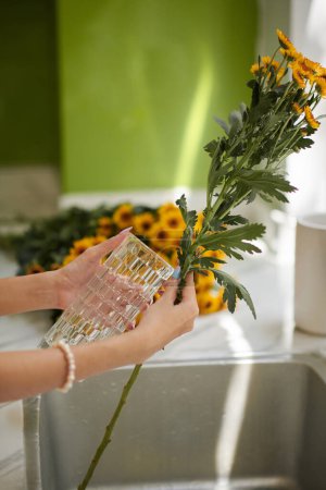 Foto de Mujer poniendo flores en jarrón de vidrio sobre fregadero de cocina - Imagen libre de derechos
