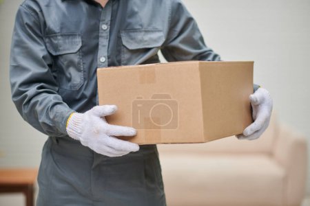 Foto de Cropped image of mover in grey uniform holding cardboard box - Imagen libre de derechos