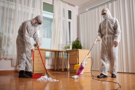 Foto de Los limpiadores en los trajes protectores que limpian y limpian al vacío el suelo en casa del cliente - Imagen libre de derechos