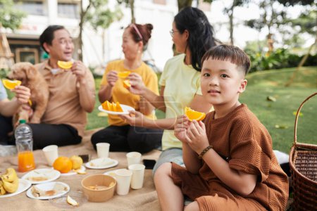 Foto de Little boy eating slice of fresh orange at family picnic - Imagen libre de derechos