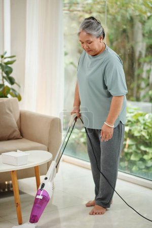 Foto de Barefoot senior woman vacuum cleaning floor in house - Imagen libre de derechos