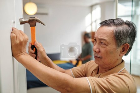 Téléchargez les photos : Senior man hammering in nails to hang picture on wall in apartment - en image libre de droit