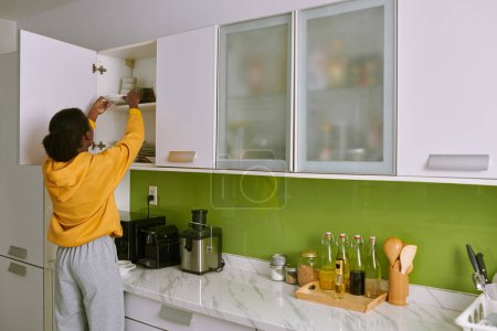Foto de Mujer en ropa de casa tomando placa fuera del gabinete de la cocina, vista desde la parte posterior - Imagen libre de derechos