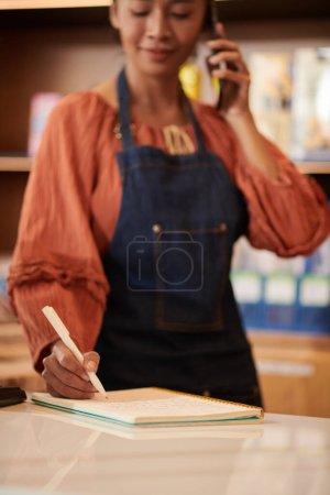 Photo for Grocery store owner ordering goods on phone and checking list in notebook - Royalty Free Image