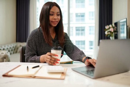 Téléchargez les photos : Femme d'affaires noire positive buvant du café à emporter du café local et travaillant sur un ordinateur portable - en image libre de droit