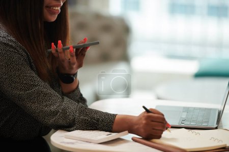 Photo for Cropped image of female entrepreneur recording voice message and taking notes in planner - Royalty Free Image