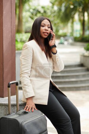 Téléchargez les photos : Portrait d'une femme d'affaires sérieuse assise à l'extérieur à côté de valises et parlant au téléphone avec un assistant - en image libre de droit