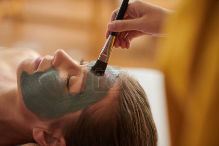 Foto de Cropped image of beautician applying purifying clay face mask with brush - Imagen libre de derechos