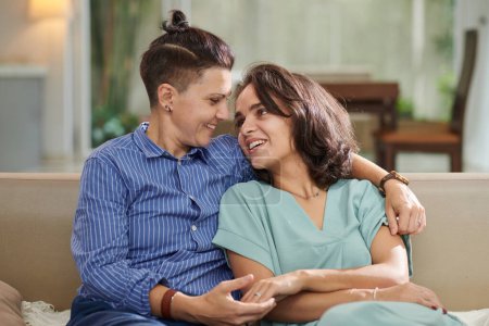 Téléchargez les photos : Portrait de couple lesbien marié assis sur un canapé à la maison et se regardant - en image libre de droit