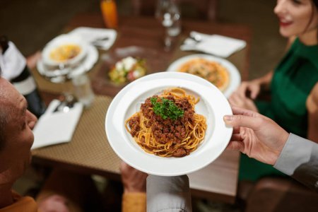 Photo for Closeup image of waiter bringing plate of spaghetti bolognese to restaurant table - Royalty Free Image