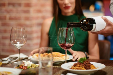 Téléchargez les photos : Serveur remplissage verre à vin pour yuong femme élégante assise à la table du restaurant - en image libre de droit